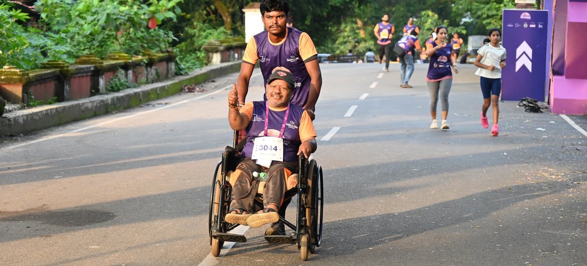 Um homem em cadeira de rodas participa de uma maratona, empurrado por um acompanhante. Outros corredores são visíveis na estrada durante o evento.