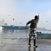 Un homme en tenue de camouflage se tient sur un quai mouillé et jette de la nourriture à une nuée de goélands. À l'arrière-plan, de grands navires sont amarrés le long de grues, sur fond de ciel nuageux.