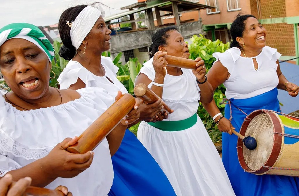 Quatre femmes afro-colombiennes en robes traditionnelles blanches et bleues jouent des maracas et du tambour lors d'une représentation culturelle.