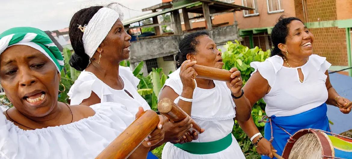 Quatre femmes afro-colombiennes en robes traditionnelles blanches et bleues jouent des maracas et du tambour lors d'une représentation culturelle.