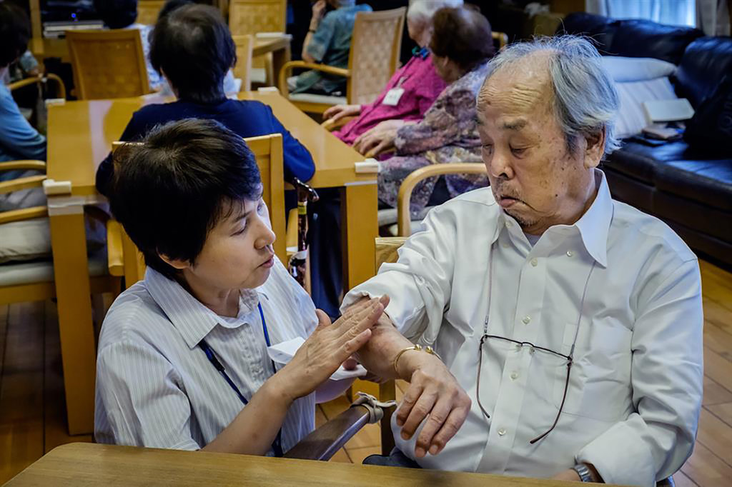 Une aide-soignante examine le bras d'un homme asiatique âgé dans ce qui semble être un établissement de soins.