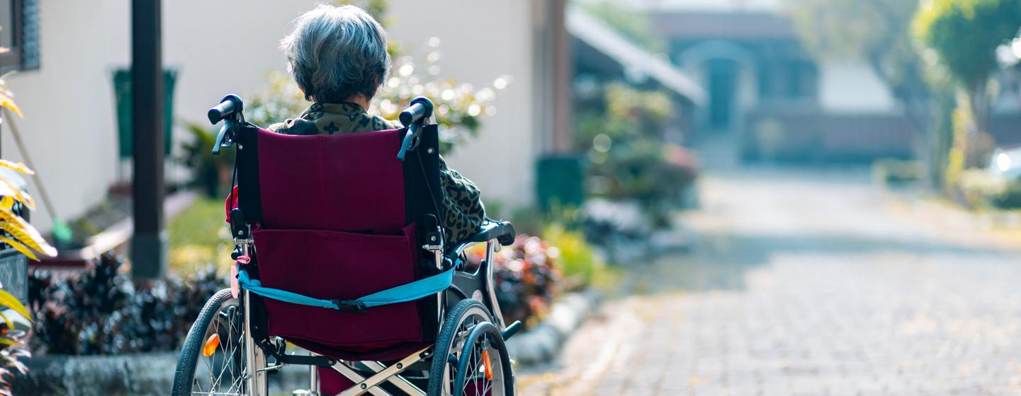 Vue de dos d'une personne âgée aux cheveux gris assise dans un fauteuil roulant bordeaux sur un chemin pavé.