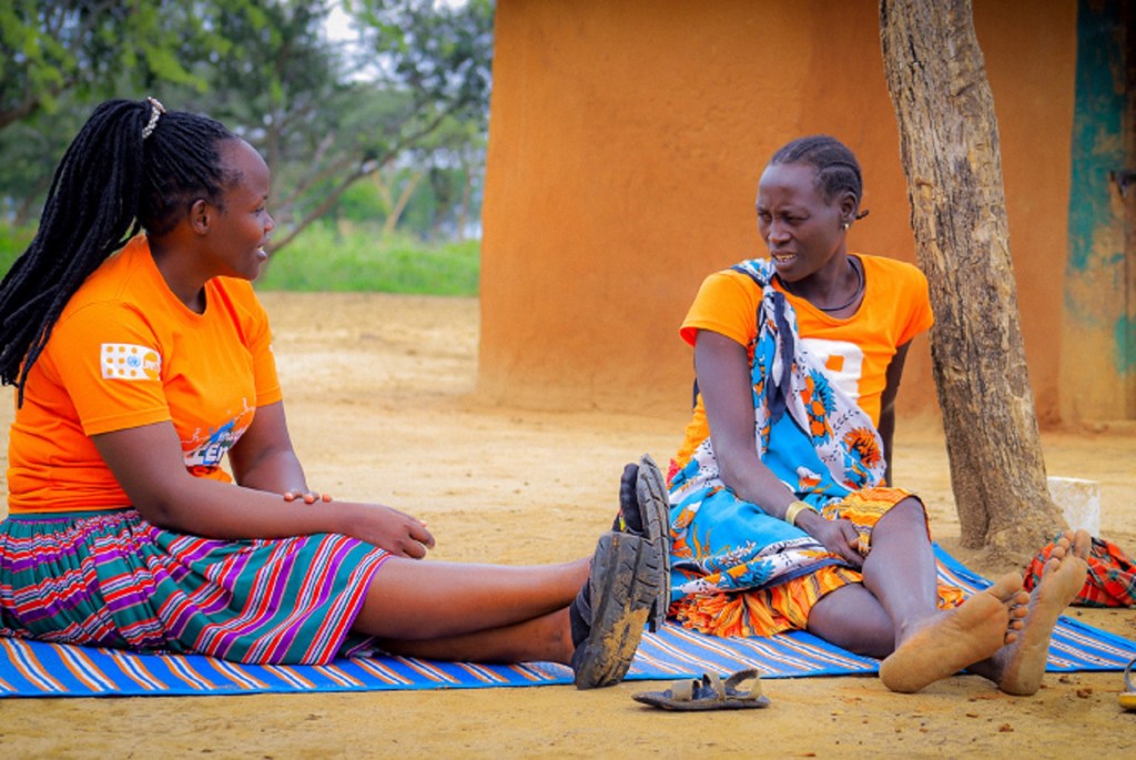 Mwanamke aliyevaa fulana ya chungwa yenye nembo ya UNFPA akizungumza na Bi Telo, ambaye pia amevalia mavazi ya chungwa. Wamekaa kwenye blanketi yenye mistari mbele ya nyumba.