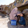 Une Afghane voilée d'une burqa administre un vaccin contre la polio à un enfant. Une glacière médicale bleue est posée à proximité, près d'un gros véhicule abandonné, dans un décor extérieur poussiéreux.