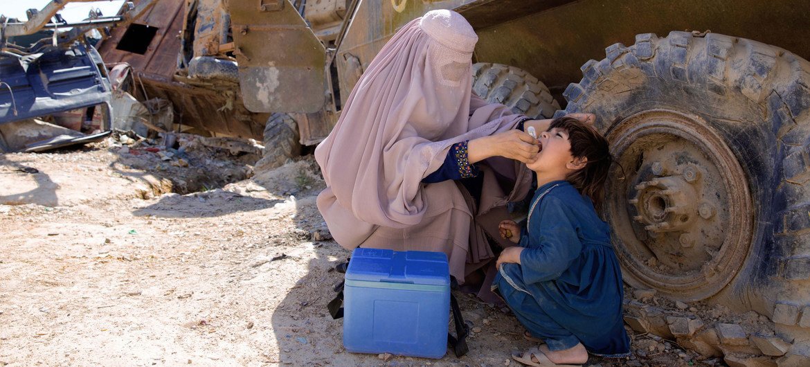 Une Afghane voilée d'une burqa administre un vaccin contre la polio à un enfant. Une glacière médicale bleue est posée à proximité, près d'un gros véhicule abandonné, dans un décor extérieur poussiéreux.
