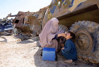 Une Afghane voilée d'une burqa administre un vaccin contre la polio à un enfant. Une glacière médicale bleue est posée à proximité, près d'un gros véhicule abandonné, dans un décor extérieur poussiéreux.