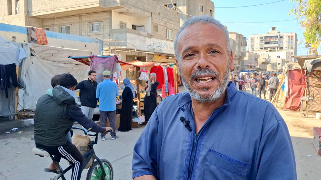 رجل ذو لحية رمادية وقميص أزرق يتحدث إلى كاميرا مزودة بميكروفون، أمام سوق خارجي صاخب في غزة.