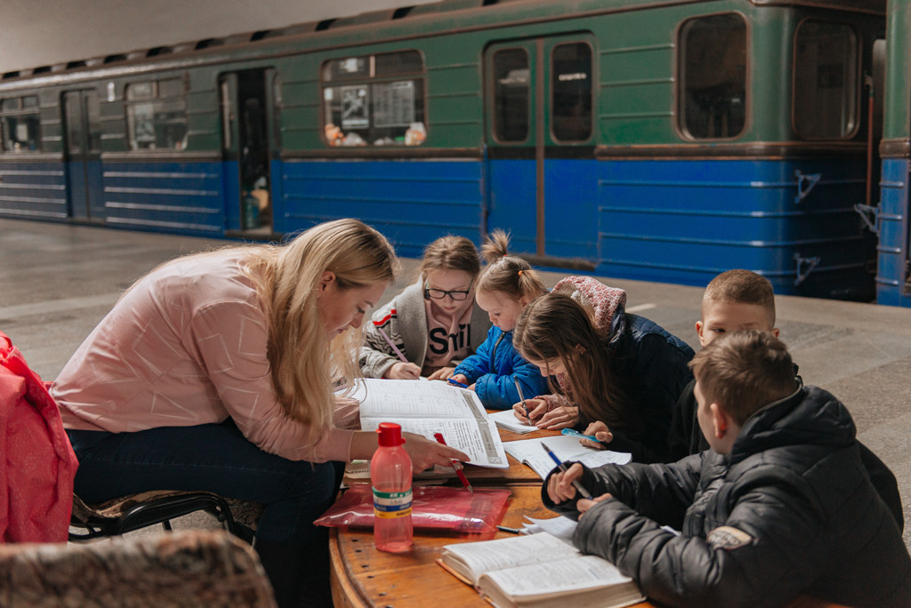 Watoto wanasomea mezani katika kituo cha treni cha Kharkiv, huku mwalimu UNICEF na watu wa kujitolea wakitoa msaada na vifaa vya kielimu.