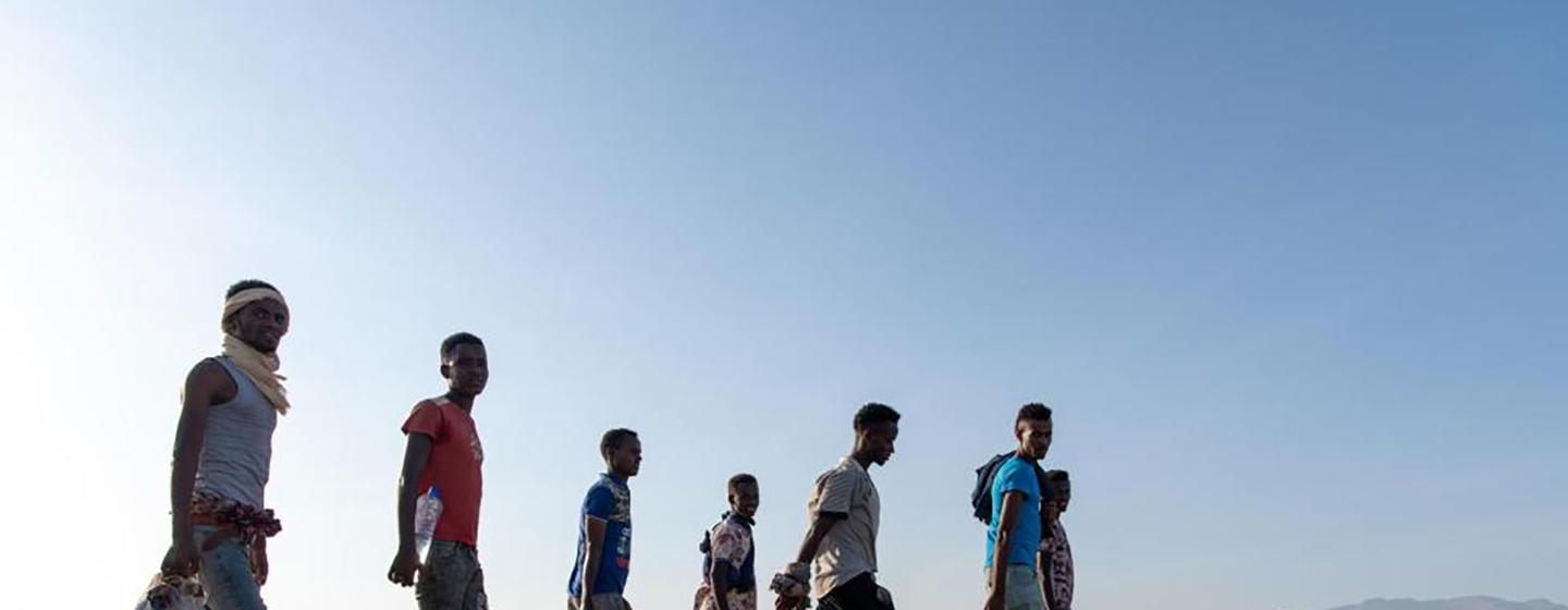 Un grupo de hombres camina a lo largo de un camino pavimentado en un paisaje seco y montañoso bajo un cielo azul claro.