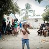 Uma menina de camisa rosa está em pé no pátio principal de um campo de deslocados internos em Léogâne, Haiti. Pessoas estão reunidas ao seu redor ao fundo.