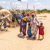 Une famille de réfugiés traverse un camp au Kenya. Un homme tient un bébé dans ses bras, tandis qu'une femme coiffée d'un foulard à motifs marche à ses côtés avec deux autres enfants. À l'arrière-plan, on aperçoit des abris de fortune et un paysage aride.
