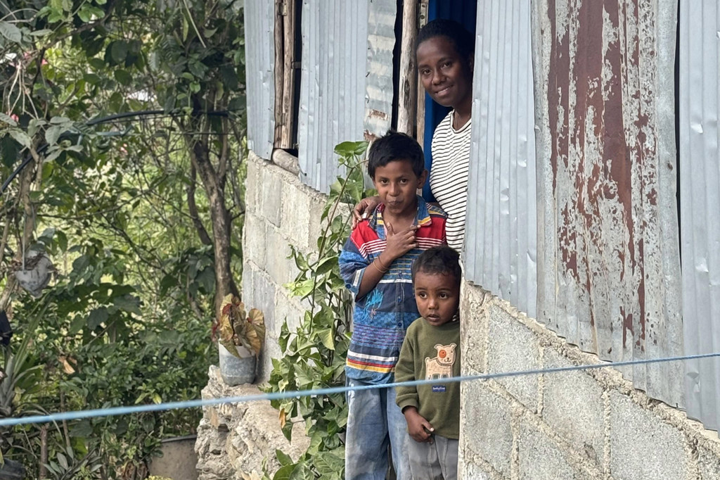 Mwanamke na wavulana wawili wadogo wamesimama mlangoni mwa nyumba ya kijijini huko Timor Leste, wakiangalia nje. Jengo hilo limetengenezwa kwa chuma cha bati na matofali ya zege, huku majani mabichi yakionekana karibu.