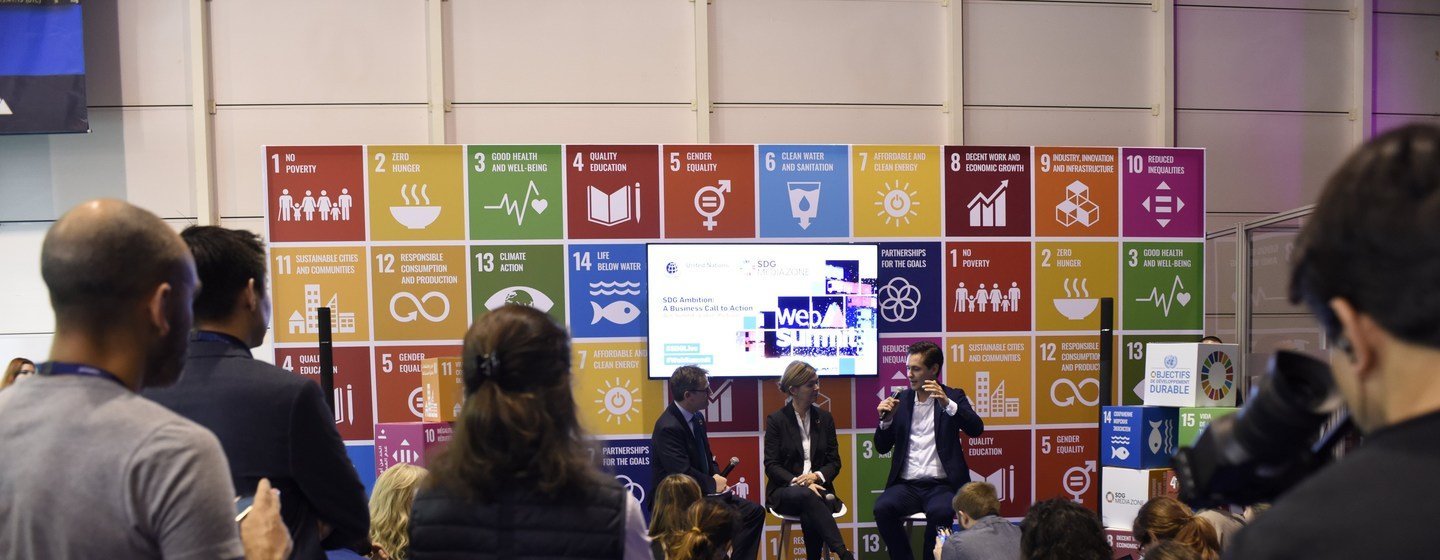 Painel de discussão no Web Summit em Lisboa, com oradores no palco em frente a um painel de fundo que exibe os Objetivos de Desenvolvimento Sustentável (ODS).