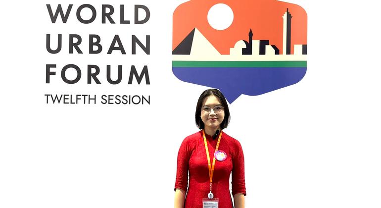 A Vietnamese woman in a red dress stands smiling in front of the logo for the Twelfth Session of the World Urban Forum.