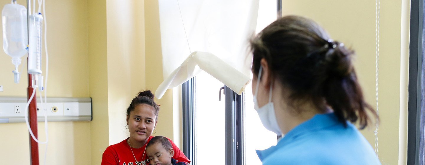 Uma funcionária da Unicef , vestindo uma camisa azul, conversa com uma mãe que segura seu filho adormecido em um quarto de hospital.