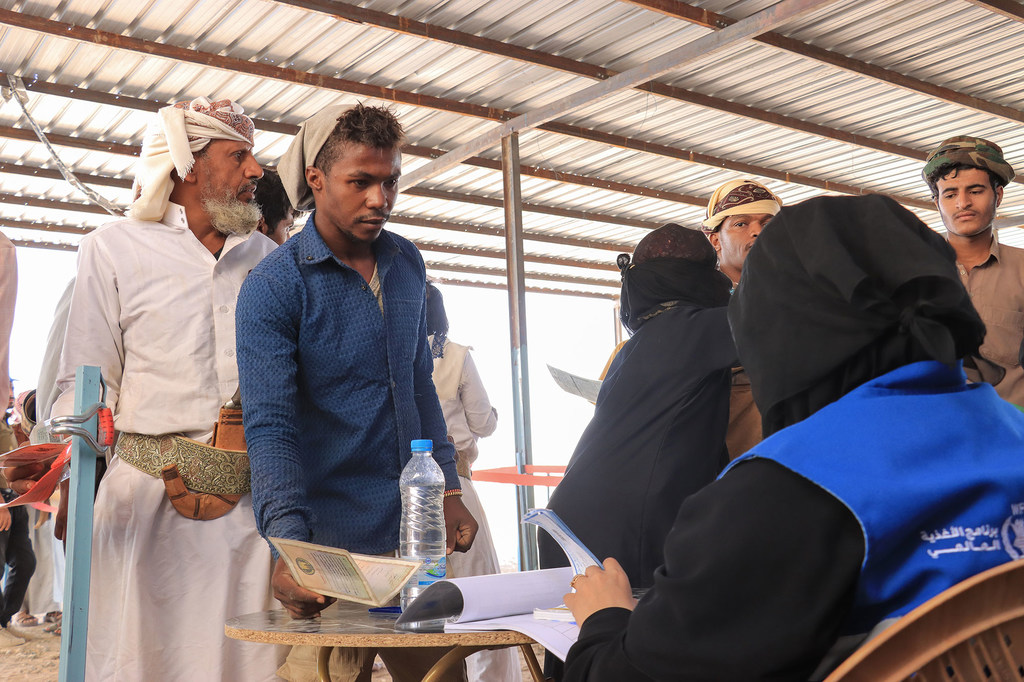 Beneficiários aguardam em fila durante um evento de distribuição de alimentos do Programa Mundial de Alimentos no Iêmen. Um trabalhador humanitário com um colete azul cadastra as pessoas em uma mesa.