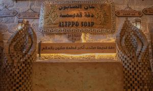 تُعرض قطعة صابون حلب التاريخية أمام جدار حجري، وعليها لافتات باللغتين العربية والإنجليزية. تُعرّف اللافتات الموقع بأنه "صناعة صابون حلب غار" و"صابون حلب"، مشيرةً إلى إدراجه في القائمة التمثيلية UNESCO للتراث الثقافي غير المادي للبشرية.