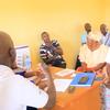 Mwanamume aliyevaa shati la WFP anapatanisha mgogoro wa kinyumbani ofisini huku wanajamii wengine wakifuatilia.