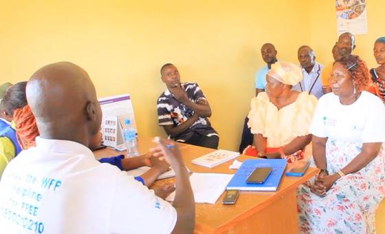 Mwanamume aliyevaa shati la WFP anapatanisha mgogoro wa kinyumbani ofisini huku wanajamii wengine wakifuatilia.