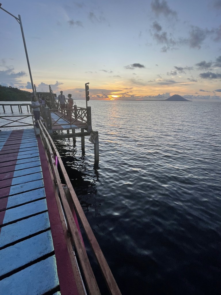 Tourists walk on a wooden pier at Budo village, Indonesia, watching the sunset over the ocean.