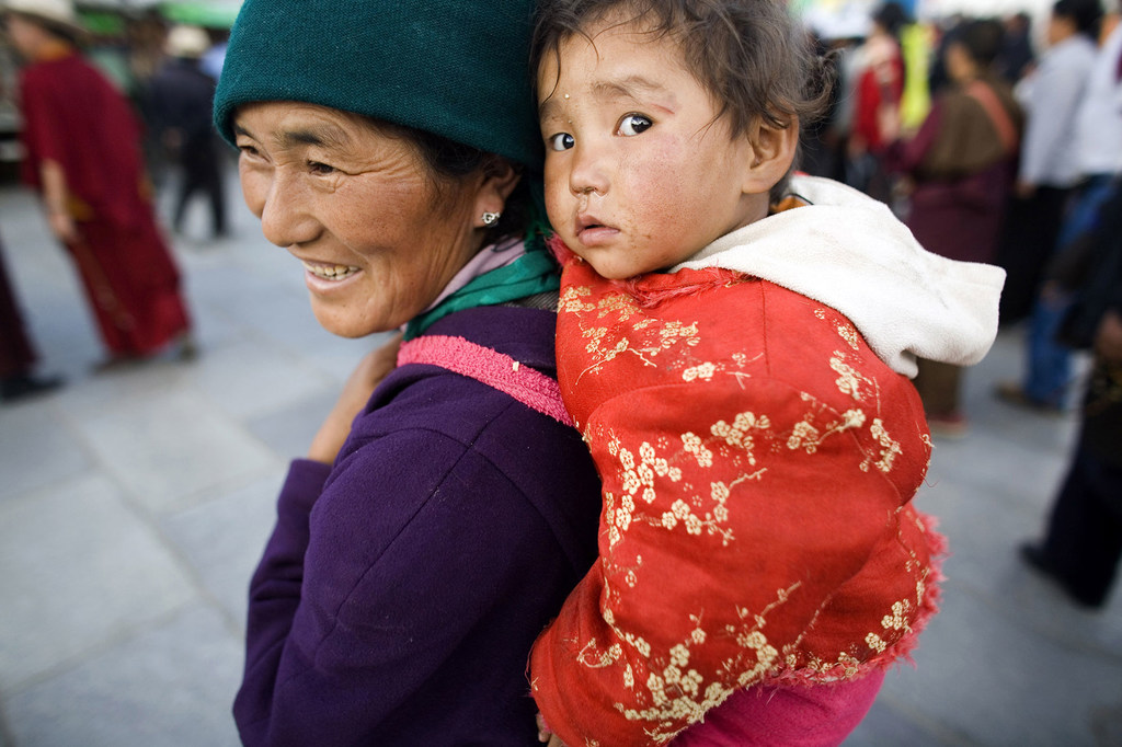 Una mujer sonriente, con sombrero verde y abrigo morado, lleva a cuestas a una niña pequeña con chaqueta roja, mientras otras personas aparecen al fondo en una calle de la ciudad.