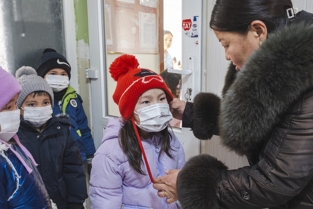 在蒙古国纳莱赫，幼儿园老师蒙赫图亚·M.正在帮助一个孩子在户外活动前戴上口罩。