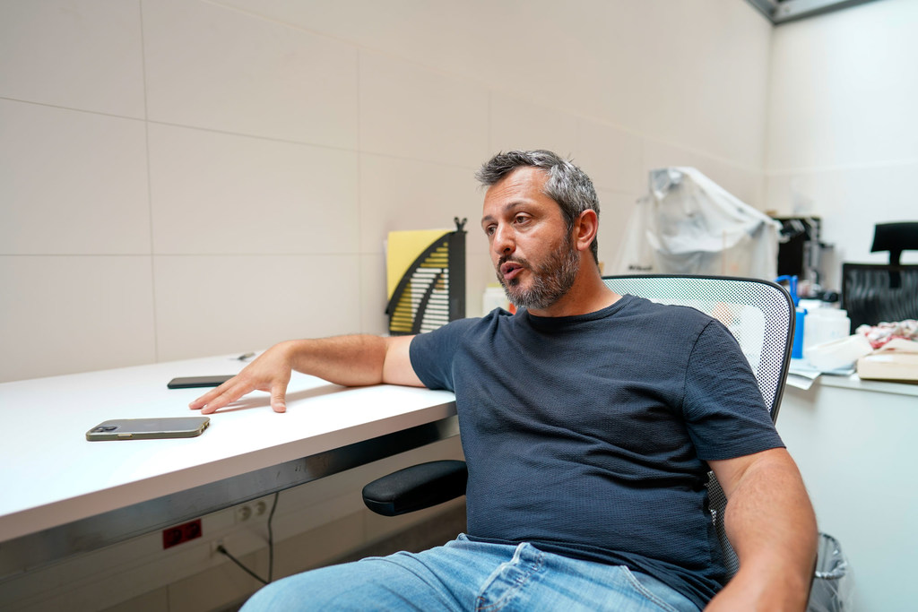 Un hombre con barba y bata de laboratorio está sentado en un escritorio en un laboratorio, mirando un teléfono inteligente.