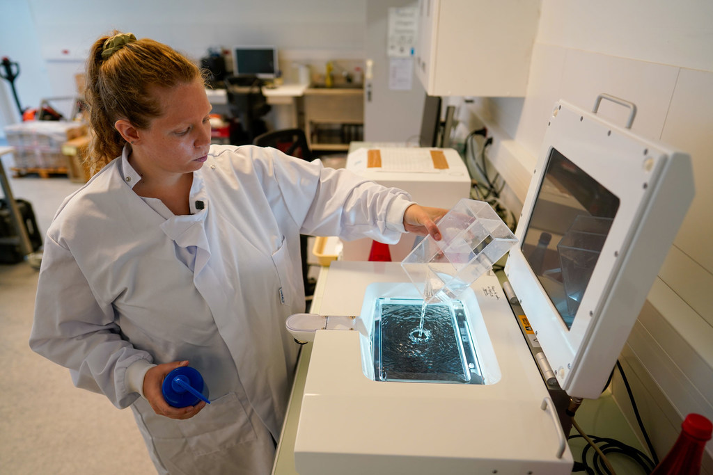 En un laboratorio, una científica vestida con una bata blanca vierte una muestra líquida de un recipiente en un equipo de laboratorio.