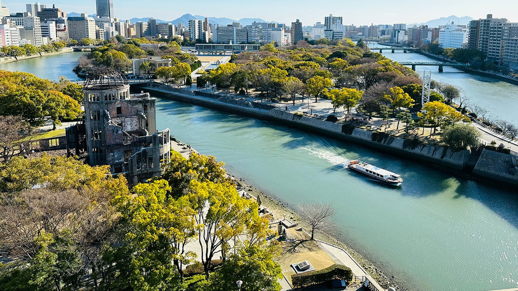 Mtazamo wa angani wa Kuba la Bomu la Atomiki na Mto Naka huko Hiroshima, Japani, ukiwa na mashua ya watalii juu ya maji na anga ya jiji la kisasa nyuma.