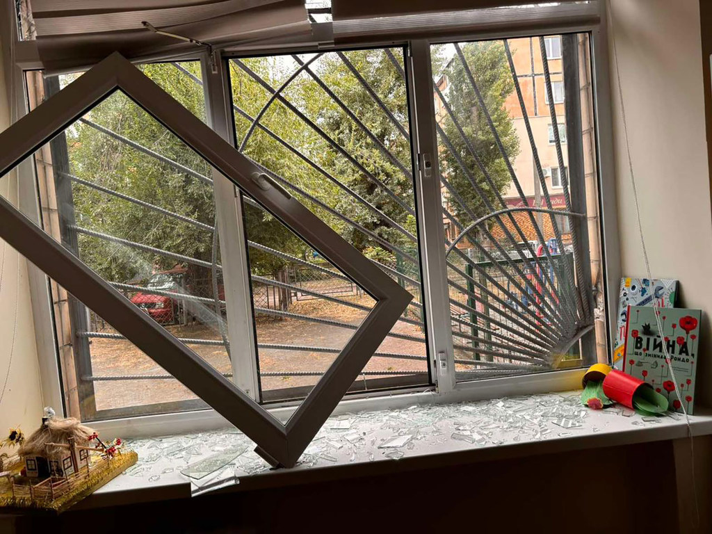 Una ventana rota en un aula de Ucrania, con cristales hechos añicos en el alféizar y libros infantiles cerca, evidencia los daños causados ​​por un ataque a un centro educativo.