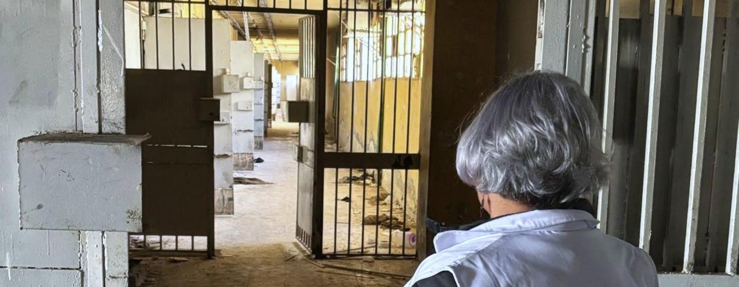 Mme Karla Quintana, directrice de l'Institution indépendante pour les personnes disparues en République arabe syrienne, vêtue d'un gilet de ONU , se tient devant une grille et regarde à l'intérieur d'un bâtiment en béton.