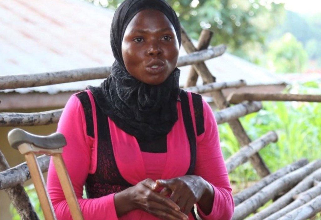 Mariama Jalloh, mkulima mwenye ulemavu nchini Sierra Leone, amesimama mbele ya uzio wa mbao akiwa na mkongojo kando yake.