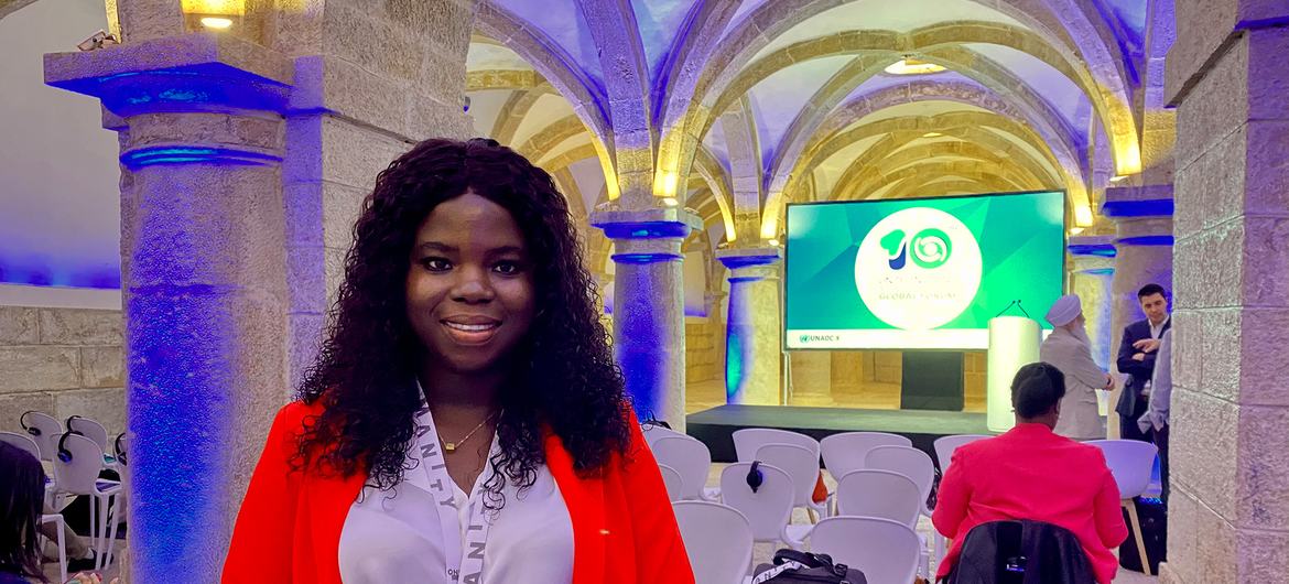 Uma mulher com jaqueta vermelha e camisa branca sorri em um salão de conferências, com um palco e um telão exibindo o logotipo do 10º aniversário da UNAOC ao fundo.