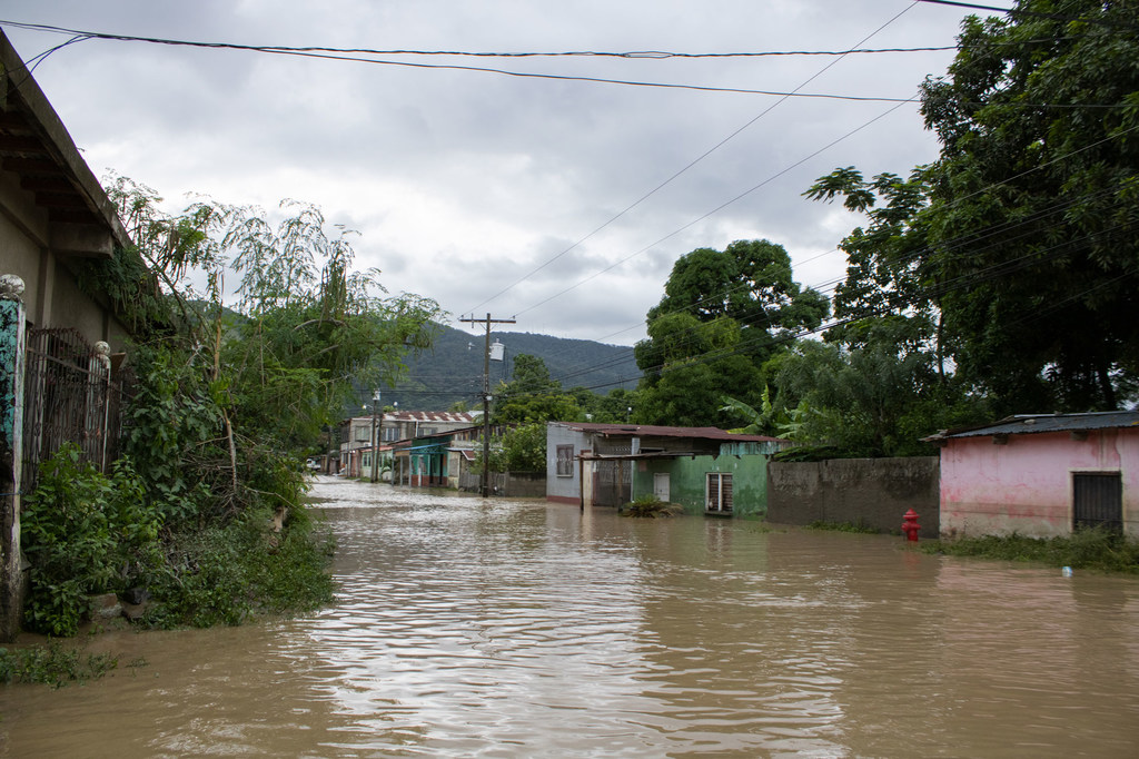 La calle principal del barrio Eben Ezer, en el sector Chamelecon de San Pedro Sula, Honduras, está inundada de agua lodosa tras la tormenta IOTA. Casas y postes de luz bordean la calle sumergida bajo un cielo nublado.