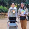 Duas mulheres em pé em uma trilha pavimentada em um parque. A mulher à esquerda, identificada como Rafaela Moreira, usa um andador. Ambas estão sorrindo e usam crachás do evento.