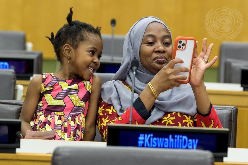 Mwanamke aliyevaa hijab na msichana mdogo wakijipiga picha ya selfie pamoja kwenye meza ya mikutano iliyoandikwa kwa ishara ya #KiswahiliDay.