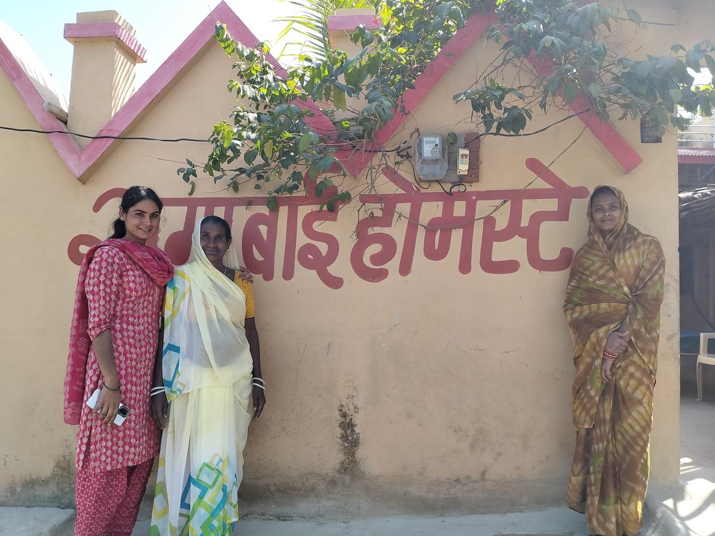 तीन महिलाएं एक इमारत के सामने पोज दे रही हैं, जिसकी दीवार पर देवनागरी लिपि में 'रामा बाई होमस्टे' लिखा हुआ है।