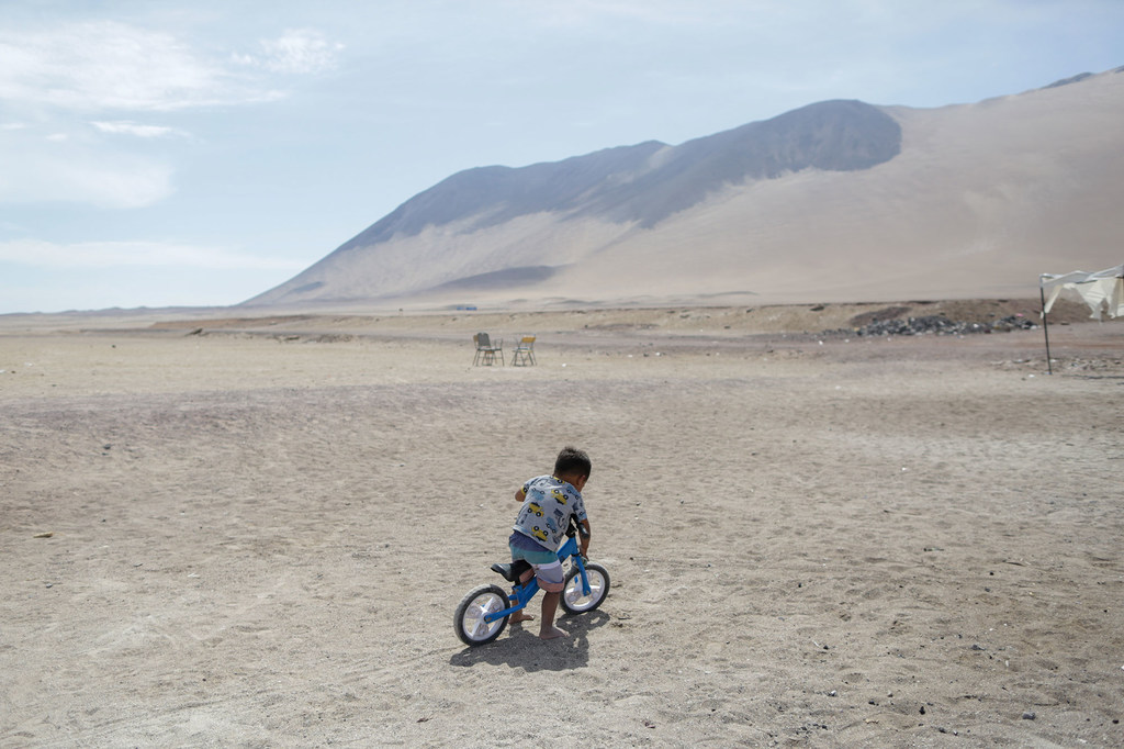 Mvulana mdogo akiendesha baiskeli ya usawa wa bluu katika eneo la jangwa katika kambi ya wahamiaji inayoungwa mkono na UNICEF huko Playa Lobitos, Chile.