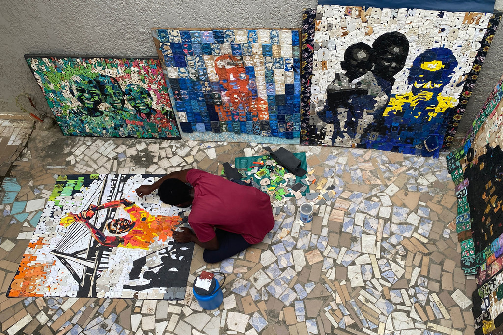 L'artiste Aristide Kouame applique des lettres découpées dans des tongs sur une toile posée au sol, tandis que d'autres mosaïques de tongs sont exposées contre le mur.