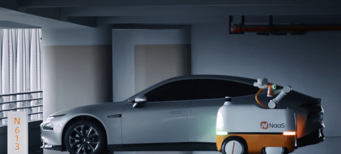 Un Tesla Model 3 plateado está estacionado junto a un robot de carga autónomo blanco y naranja con el logotipo de NaaS en un estacionamiento.