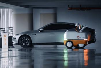 Un Tesla Model 3 plateado está estacionado junto a un robot de carga autónomo blanco y naranja con el logotipo de NaaS en un estacionamiento.