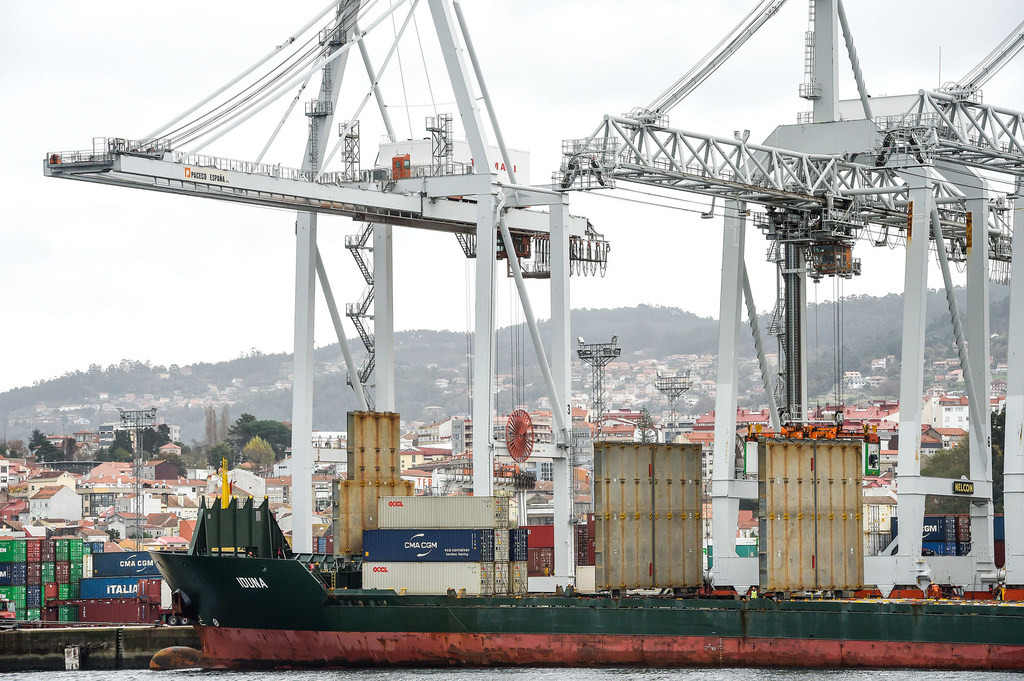 Un buque de carga con contenedores está amarrado en el puerto comercial de Vigo, España, con grandes grúas y una ciudad costera al fondo.