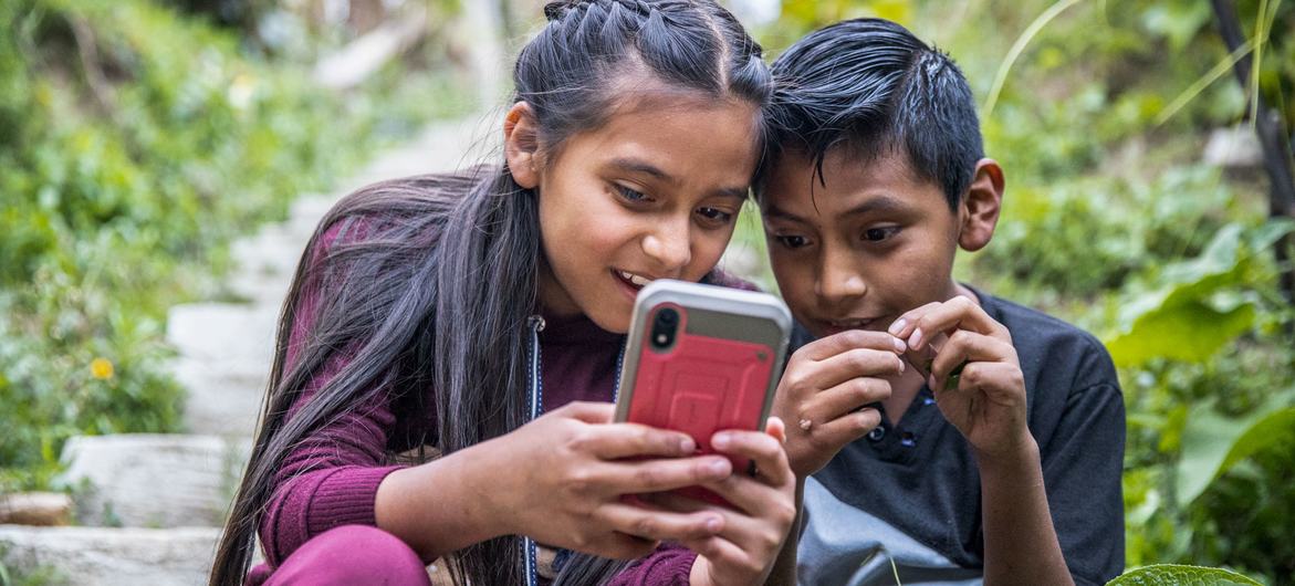 Dos niños juegan con su teléfono al aire libre mientras sus padres participan en un taller sobre seguridad en la red y prácticas parentales positivas, en Guatemala.