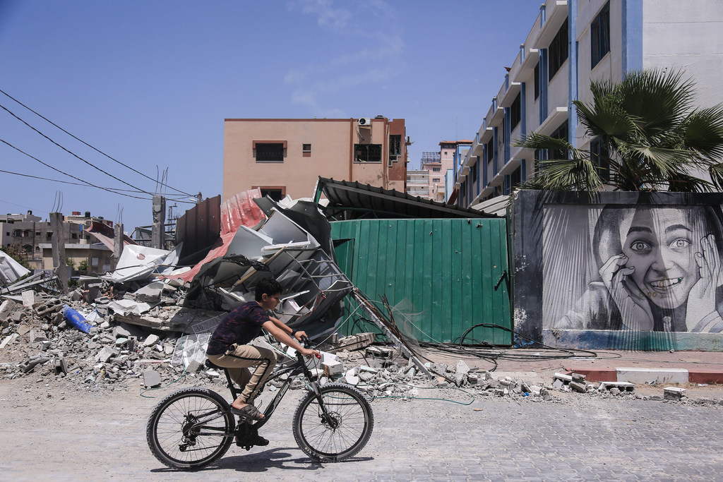 Un niño palestino monta en bicicleta por una calle llena de escombros de edificios destruidos en Gaza, con un mural en una pared al fondo.