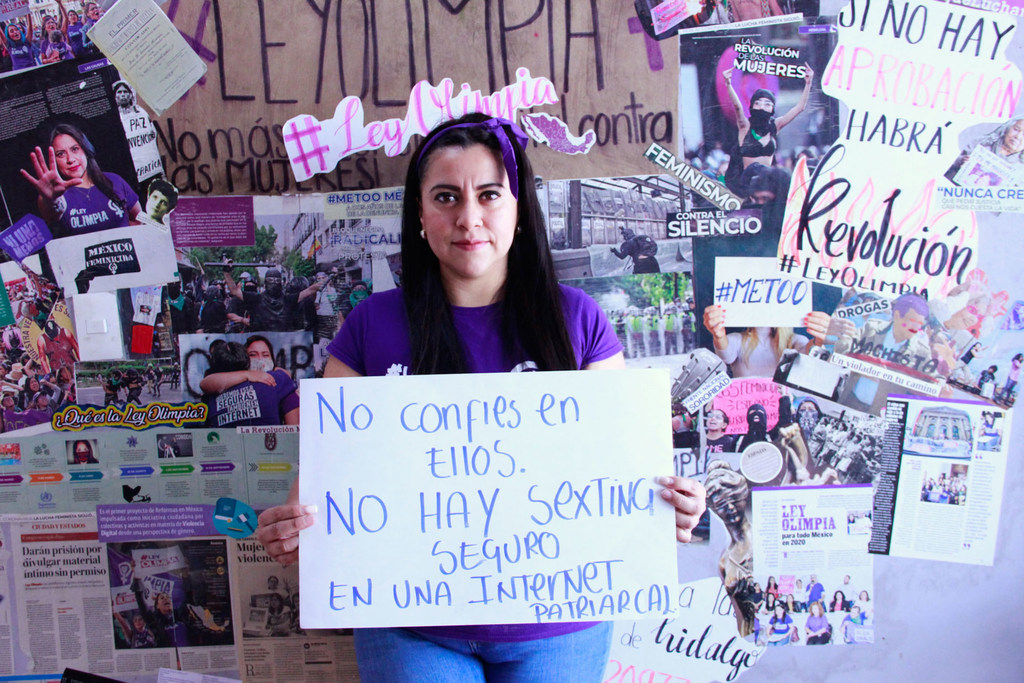 Una activista con cabello largo y oscuro y una diadema morada sostiene un cartel blanco de protesta. El cartel dice: «No confíes en ellos. NO HAY SEXTING SEGURO EN UNA INTERNET PATRIARCAL». Se encuentra frente a un muro cubierto de carteles y volantes de protesta relacionados con la Ley Olimpia y el movimiento #MeToo en México.