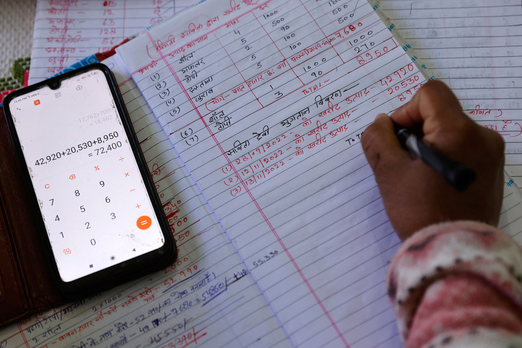एक नोटबुक में हाथ से की गई गणनाओं के बगल में एक स्मार्टफोन कैलकुलेटर रखा है, जिसमें 72,400 का योग दर्शाया गया है।