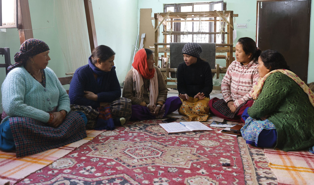 पारंपरिक पोशाक पहने पांच महिलाएं एक कार्यशाला में एक पैटर्न वाले कालीन पर बैठी हैं, जिसके पृष्ठभूमि में हथकरघा बुनाई के फ्रेम दिखाई दे रहे हैं।