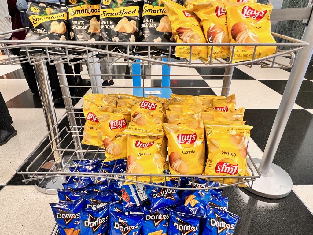 Um carrinho expositor de arame metálico repleto de pacotes de salgadinhos Lays, Doritos e Smartfood em uma loja com piso quadriculado em preto e branco.
