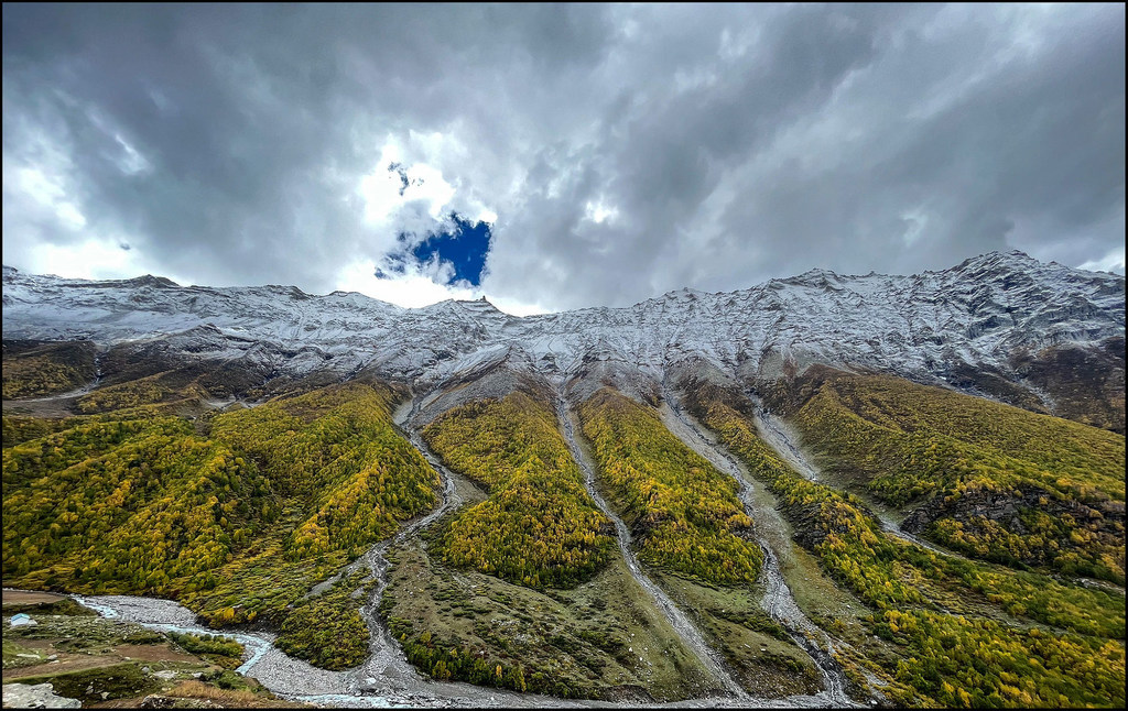 Una toma panorámica de montañas nevadas con follaje otoñal verde y amarillo en las laderas inferiores y un río que serpentea a través de un valle en el Himalaya, India.