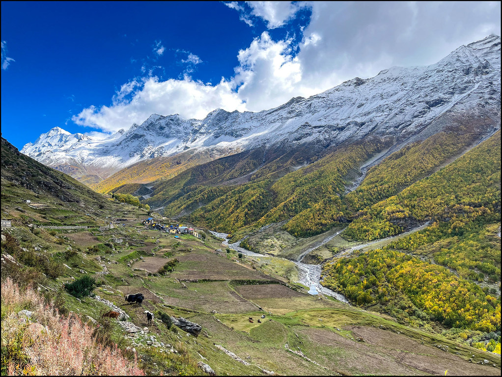हिमालय की बर्फ से ढकी पर्वत चोटियों से घिरी एक घाटी दिखाई देती है, जिसमें सीढ़ीदार खेत, एक नदी और बादलों से भरे नीले आकाश के नीचे एक छोटा सा गांव बसा हुआ है।